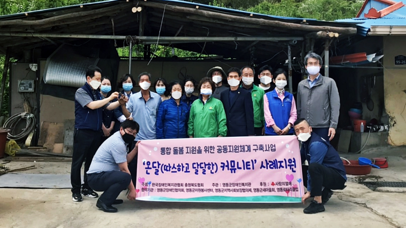 통합돌봄 지원을 위한 공동지원체계 구축사업의 하나로 사례지원을 한 가정에서 후원자와 대상자분들의 기념 촬영을 한 사진입니다. 사진의 내용은 14명이 서 있고 앞에 두명은 앞에 앉아서 온달커뮤니티 사례지원 플랜카드를 앉은 자세로 양쪽으로 들고 있는 사진입니다.